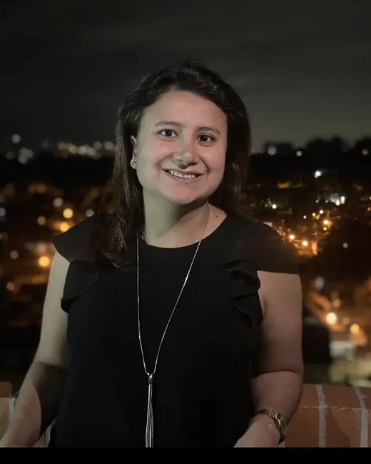 Miss Mari sonriendo de noche, con la ciudad iluminada al fondo